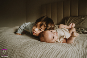 Brother Presses Gently Beside Baby Sister In Peaceful And Tenderly Lit Bedroom Portrait In Toronto In this softly lit Toronto bedroom, a young brother presses close beside his baby sister with effortless tenderness. The warm shadows and low perspective make the photograph feel intimate, peaceful, and beautifully centered on quiet sibling connection.