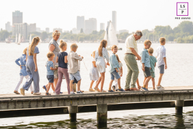 Grandparents Lead Parade Of Grandchildren Beside Water On Dock At Kralingse Plas In Rotterdam Along the dock at Kralingse Plas in Rotterdam, grandparents lead a parade of grandchildren beside the water, each child adding their own rhythm to the scene. The layered line of figures and distant skyline give the image wonderful energy and connection.