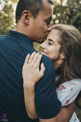 In Mississauga, Ontario, the engaged couple shares a quiet embrace as he kisses her forehead and she smiles against his shoulder. The close framing and soft natural light give the portrait intimacy, tenderness, and genuine affection.