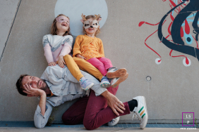 Exhausted Paris Father Is Human Bench as Girls Burst Into Laughter in Honest Family Portrait Theh exhausted Paris father becomes a human bench while the girls burst into laughter above him, completely delighted by the arrangement. The playful body language and whimsical wall art give the portrait humor, chaos, and wonderfully honest family energy