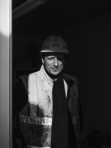 Um retrato empresarial em preto e branco de um trabalhador da construção da Spie Batignolles Fondations, usando um capacete, descansando em um canteiro de obras em Val de Marne. A luz do sol projeta sombras dramáticas em seu rosto.