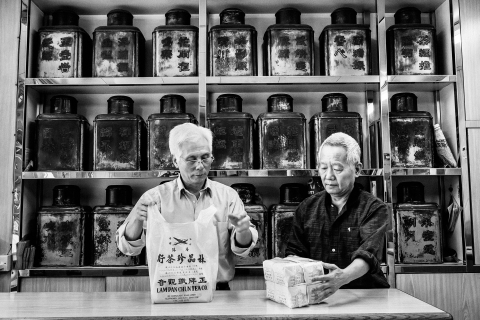 Retrato profissional de negócios de dois homens da Lam Pan Chun Tea Company em Hong Kong, capturados em uma impressionante foto em preto e branco enquanto ensacam diligentemente os pedidos. Esta imagem mostra a natureza trabalhadora e dedicada da empresa.