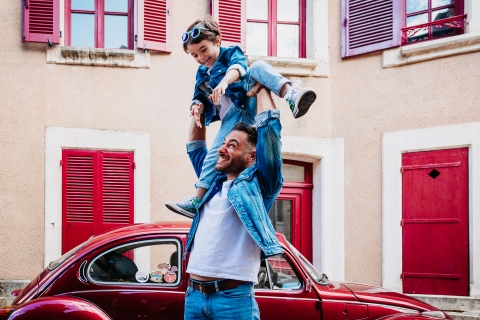 An Auvergne-Rhone-Alpes dad is playing with his boy near a red car and a building with red doors, windows and shutters