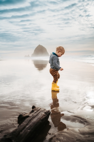 An adorable toddler with boundless curiosity wanders the enchanting sands of Cannon Beach, Oregon, during low tide, donning precious little yellow boots