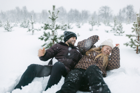 In a cozy scene from Lofoten, Norway, a couple sits together in the snow, playfully tossing snow and laughing in the midst of towering pine trees.