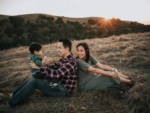 Uma foto foi tirada no Ed R Levin County Park mostrando mamãe e papai sentados de costas um para o outro na grama, com papai segurando a criança e o pôr do sol brilhando sobre as colinas.