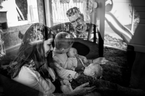 A imagem mostra uma família aproveitando o tempo junta em casa em Montpellier, França, com foco em um pai segurando um bebê, capturado em preto e branco através de um vidro.