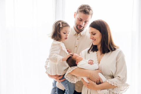 Sessão de retratos de estilo de vida em Lyon, França, capturando o vínculo amoroso entre uma família de quatro pessoas. A irmã mais velha protege e se comunica com sua irmã recém-nascida nos braços de seus pais.