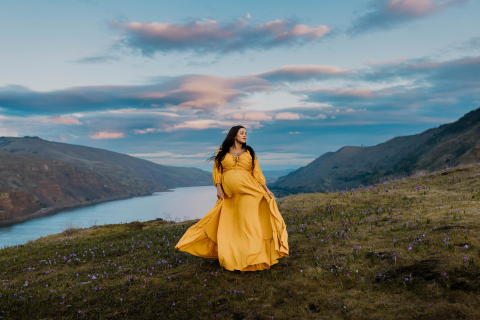 Em Hood River, Oregon, uma mãe grávida está graciosamente nas montanhas, usando um vestido amarelo esvoaçante que contrasta lindamente com a paisagem natural acidentada e a vegetação vibrante ao seu redor.