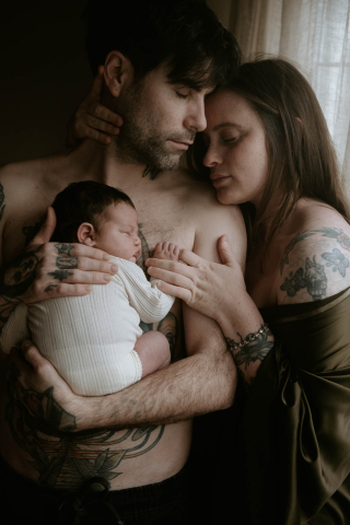 Em Margate, Inglaterra, uma família de três pessoas compartilha um momento terno em casa. O casal segura amorosamente seu recém-nascido com os olhos fechados, saboreando a intimidade silenciosa e o calor de sua nova vida juntos.