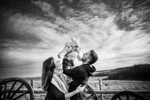 Em Hérault, uma alegre fotografia de família captura pais alegremente levantando seu bebê no ar, todos sorridentes, com o cenário pitoresco de uma paisagem ensolarada e cênica acrescentando calor e alegria ao momento.