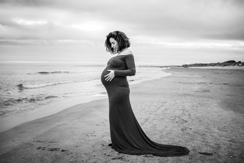 Em Hérault, uma fotografia de maternidade captura uma radiante mãe grávida parada na praia, usando um lindo vestido de maternidade que flui suavemente na brisa do mar, com as ondas batendo suavemente na costa atrás