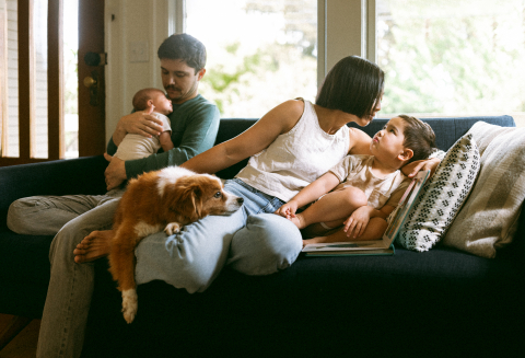 Um lindo retrato de família em Portland, Oregon. O pai segura seu recém-nascido no sofá enquanto a mãe conversa com seu filho pequeno e um cachorrinho senta em seu colo.