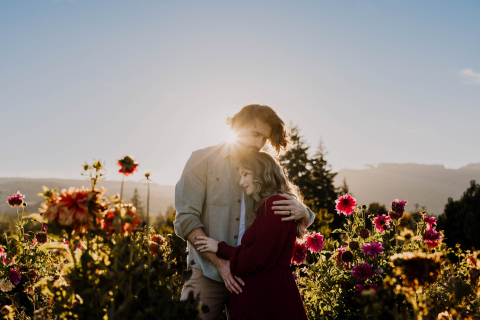 Um retrato de estilo de vida capturado em Hood River, Oregon. Um casal grávido em pé em um campo de flores iluminado pelo sol, seu amor irradiando enquanto eles se abraçam com alegria e antecipação. O raio de sol atrás de sua cabeça adiciona um toque a esta cena ao ar livre.