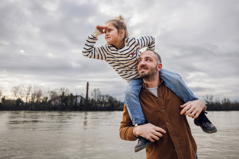 Em Lyon, França, um pai carrega a filha nos ombros enquanto ela se inclina para a frente, protegendo os olhos com a mão, e olha para o longe.