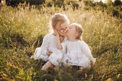 Em Lyon, França, duas irmãs estão sentadas na grama, com a mais nova beijando docemente a mais velha na bochecha, banhadas pela luz do sol em meio à beleza da natureza.