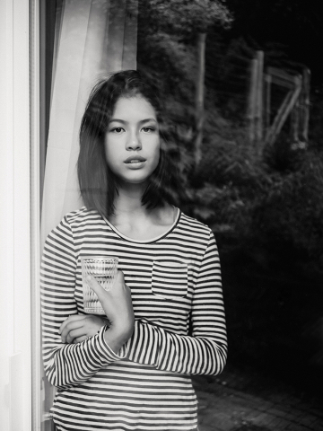 Em Braeside, Ontário, Canadá, uma menina é capturada em preto e branco em uma manhã de início de verão, em pé em frente a uma janela. A imagem reflete um reflexo tranquilo e uma luz natural suave.