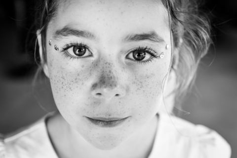 Em Vanzy, Alta Saboia, França, um retrato em preto e branco captura uma jovem sorrindo, destacando seus olhos cativantes e sardas. A foto íntima reflete sua beleza natural e espírito alegre.