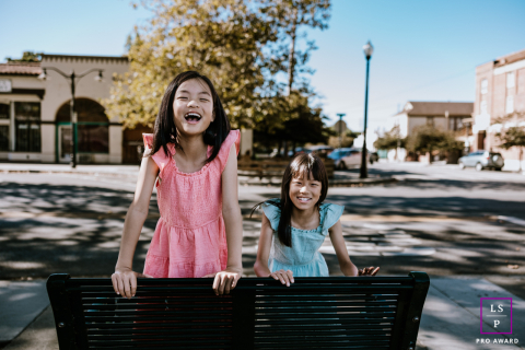 Duas meninas, gêmeas, sentam-se em um banco no coração de Fremont, Califórnia. Uma está vestida em tons de verão, enquanto a outra incorpora o calor e os tons do outono. Enquanto elas se ajoelham juntas, seus vestidos de cores contrastantes criam um contraste visual.