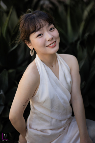 Capturado em Santa Clara, Califórnia, este retrato de estilo de vida irradia beleza e tranquilidade. A mulher está sozinha, adornada com um vestido branco simples, seu sorriso gentil direcionado para a câmera. 