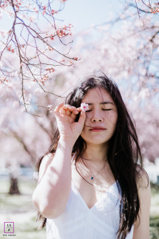 Jovem abraça a serenidade da primavera sob árvores floridas em San Ramon, Califórnia Em San Ramon, Califórnia, uma jovem está sob as flores da primavera, de olhos fechados, segurando delicadamente uma única flor. Ela aprecia a serenidade da paisagem, cercada pela beleza vibrante da estação.