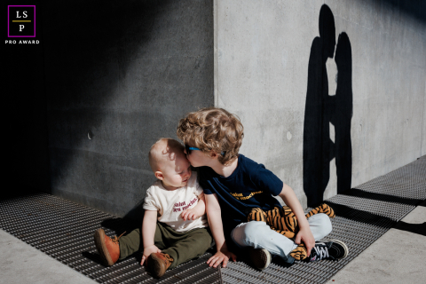 Em Lyon, França, crianças sentam-se e compartilham um beijo, enquanto as sombras dos pais se beijando são projetadas no muro de concreto urbano atrás delas.
