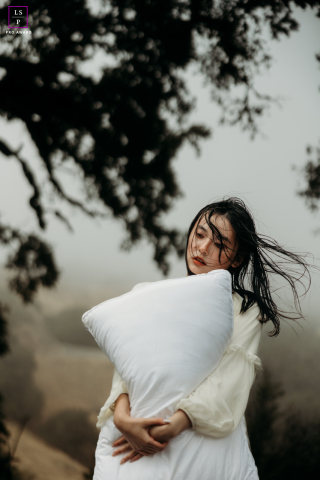 Soft Sun, San Bruno: Mulher sonhadora abraça seu travesseiro Em San Bruno, Califórnia, uma mulher está sob as árvores, perdida em devaneios. O vento sopra suavemente seus cabelos enquanto ela abraça ternamente um travesseiro, enquanto a suave luz do sol se filtra pelas folhas.