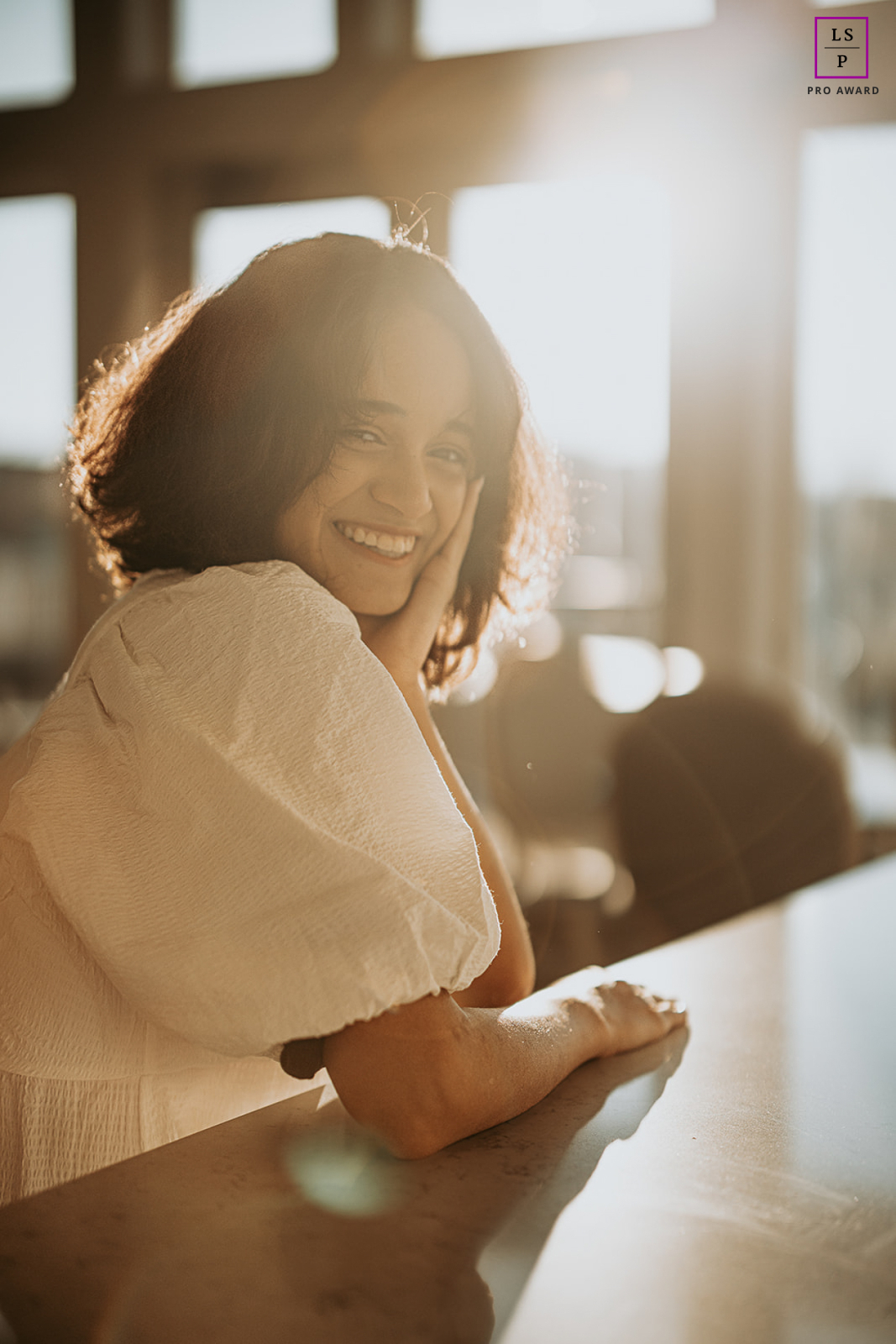 Capturada em Champaign, Illinois, uma jovem recém-formada irradia pura felicidade enquanto sorri ao sol, sentada em uma mesa alta em um ambiente interno próximo a grandes janelas.
