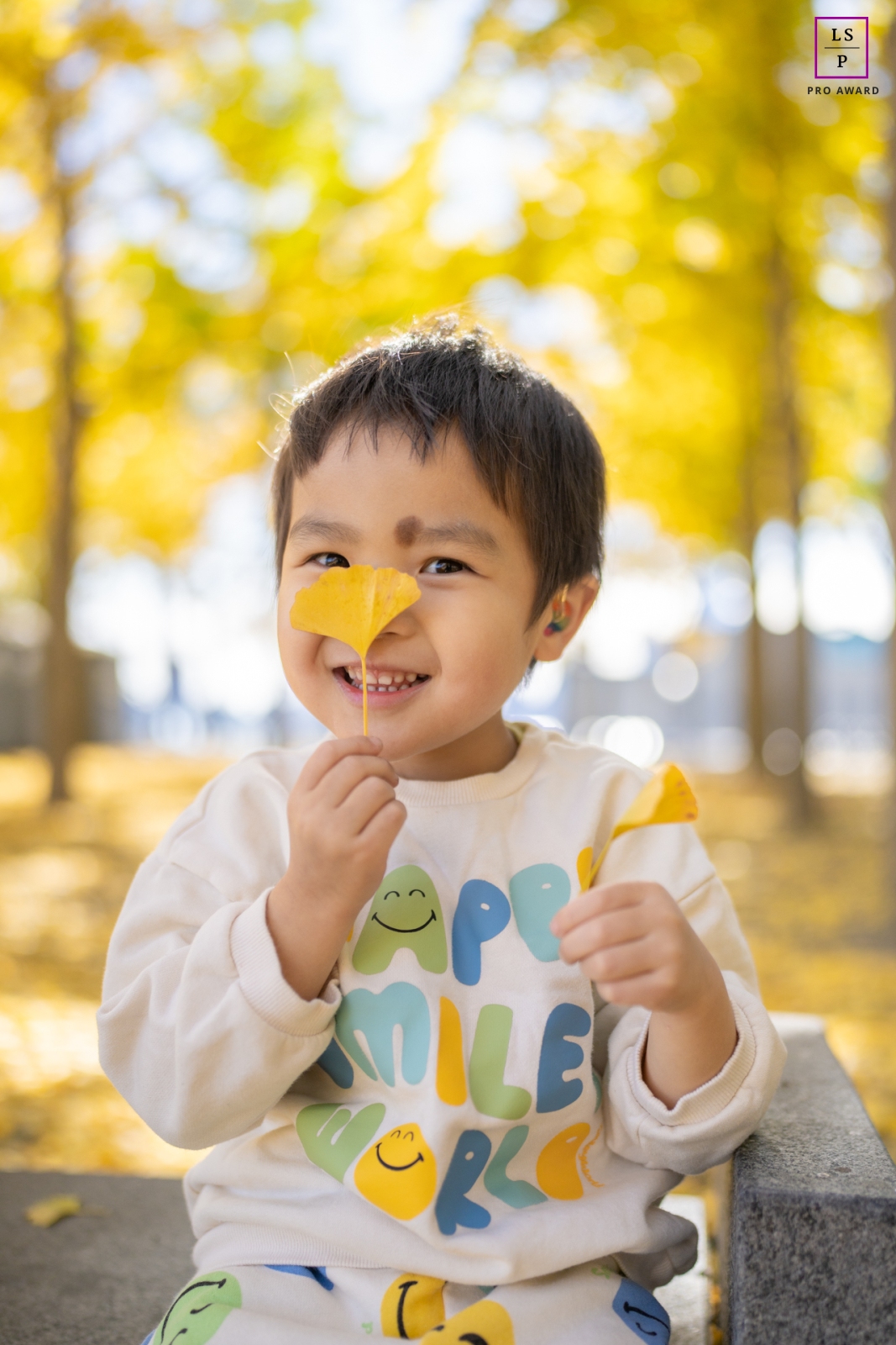 Um retrato tirado no lindo cenário de um parque em Hoboken, NJ. Em primeiro plano, um jovem rapaz com um sorriso segura uma folha amarela vibrante até o nariz, escondendo-se atrás dela de forma divertida. O cenário mostra as cores deslumbrantes do outono nas árvores.