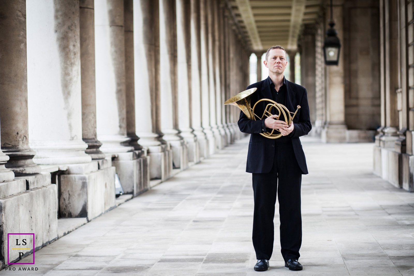 A stylish man in London, England, confidently poses with a French horn, framed by imposing pillars.