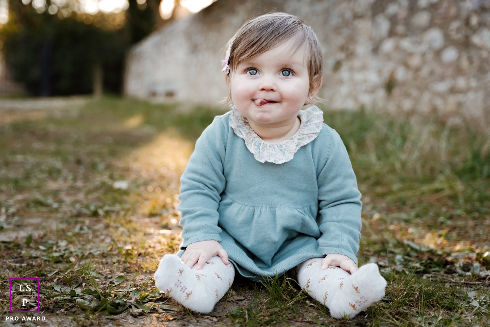 This adorable child is enjoying the sunshine in Montpellier, smiling with their tongue sticking out in a cute pose while sitting on the short grass