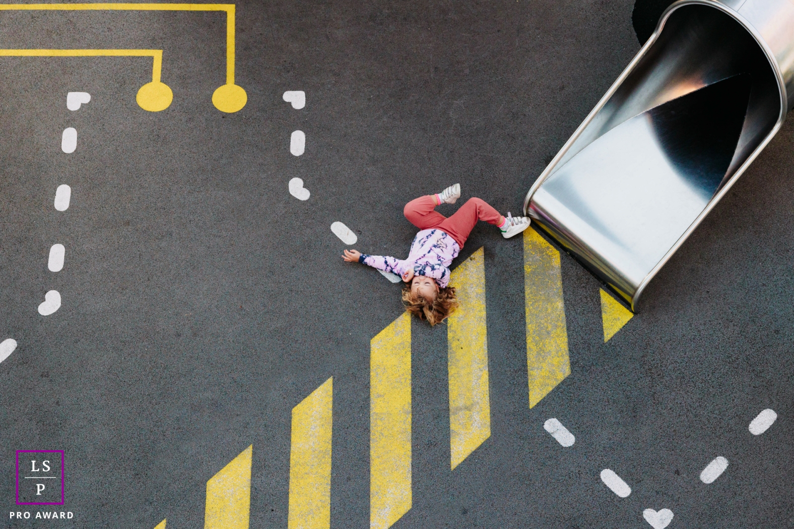 In a vibrant New York playground, captured from above, a girl gracefully descends from a slide, finally landing and laying on the beautifully painted floor, showcasing her playful lifestyle
