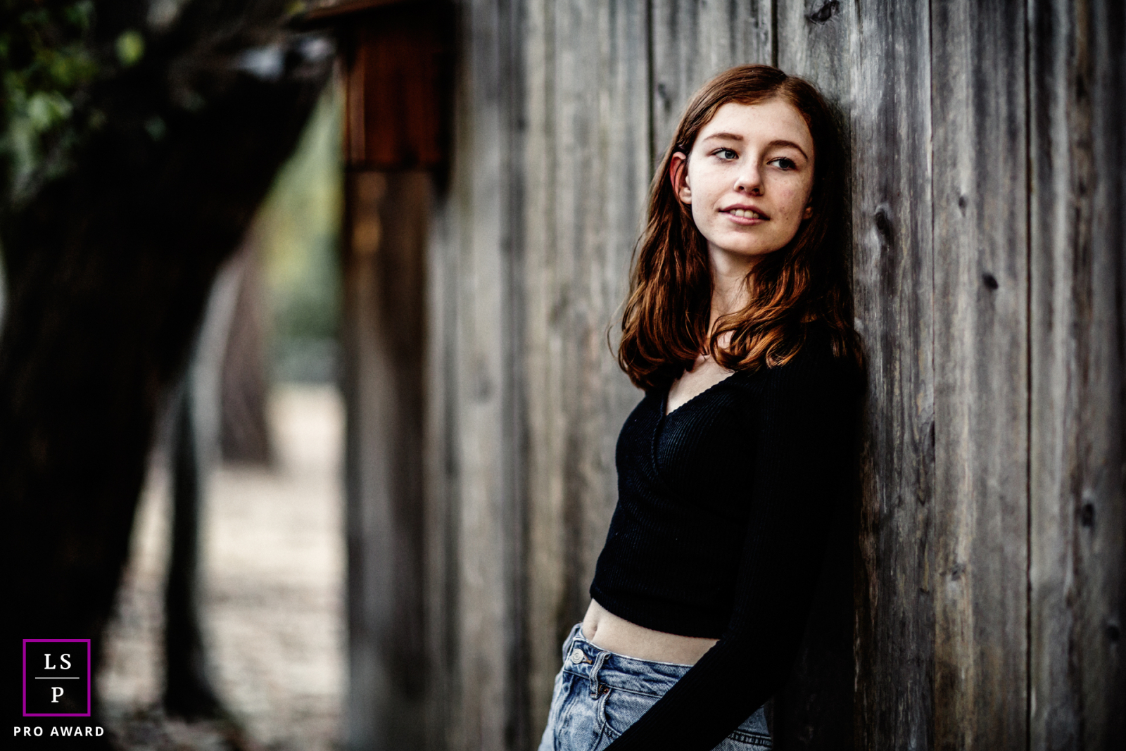 Um retrato de estilo de vida tirado em Los Altos, CA, apresentando uma jovem mulher encostada em uma cerca. Ela olha para longe, suas costas e cabeça confortavelmente apoiadas na estrutura de madeira.