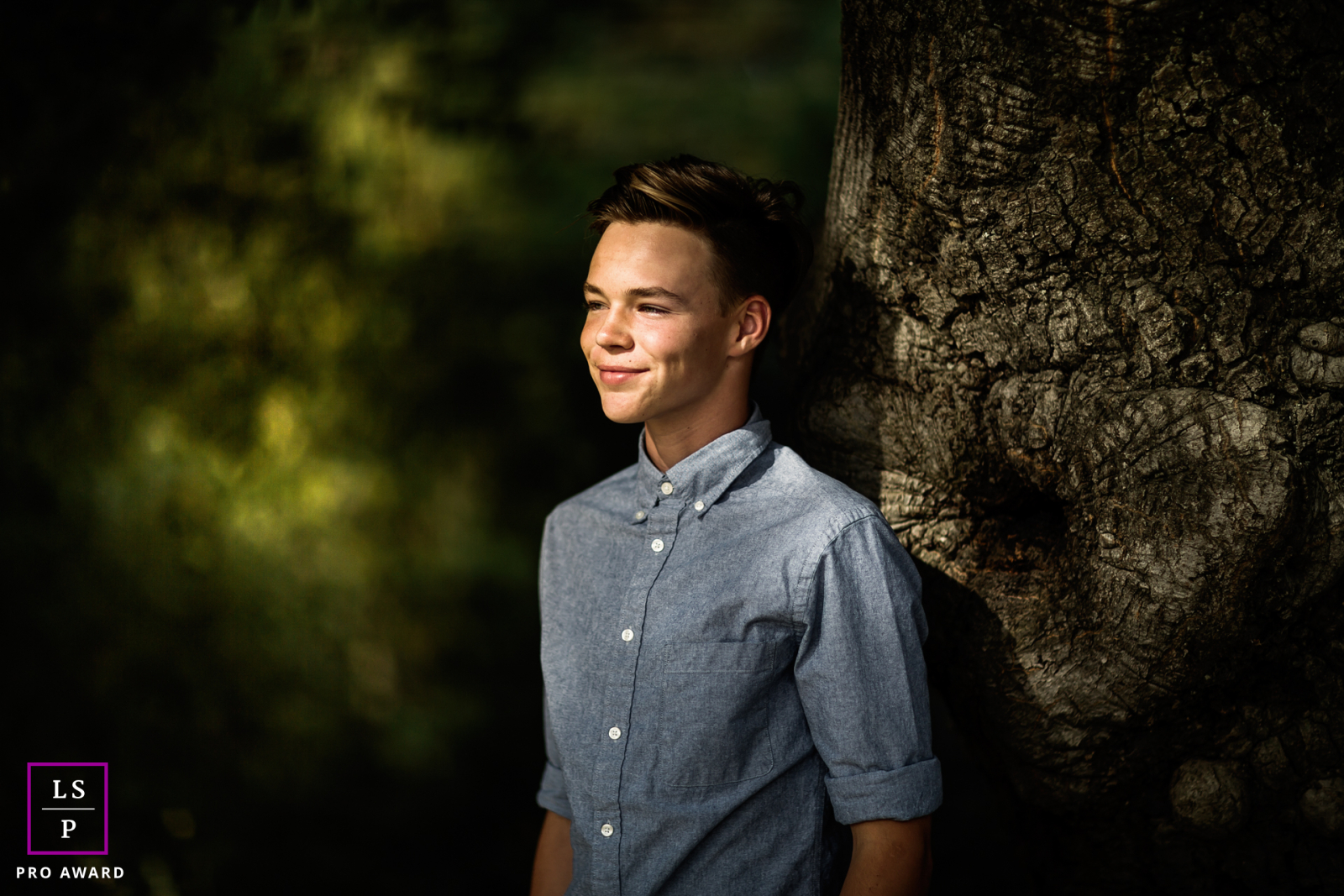 Um retrato em Cupertino, CA mostra um jovem adolescente parado ao lado de um tronco de árvore, iluminado pela luz do sol salpicada que filtra através das árvores. Ele olha para longe com um sorriso brilhante.
