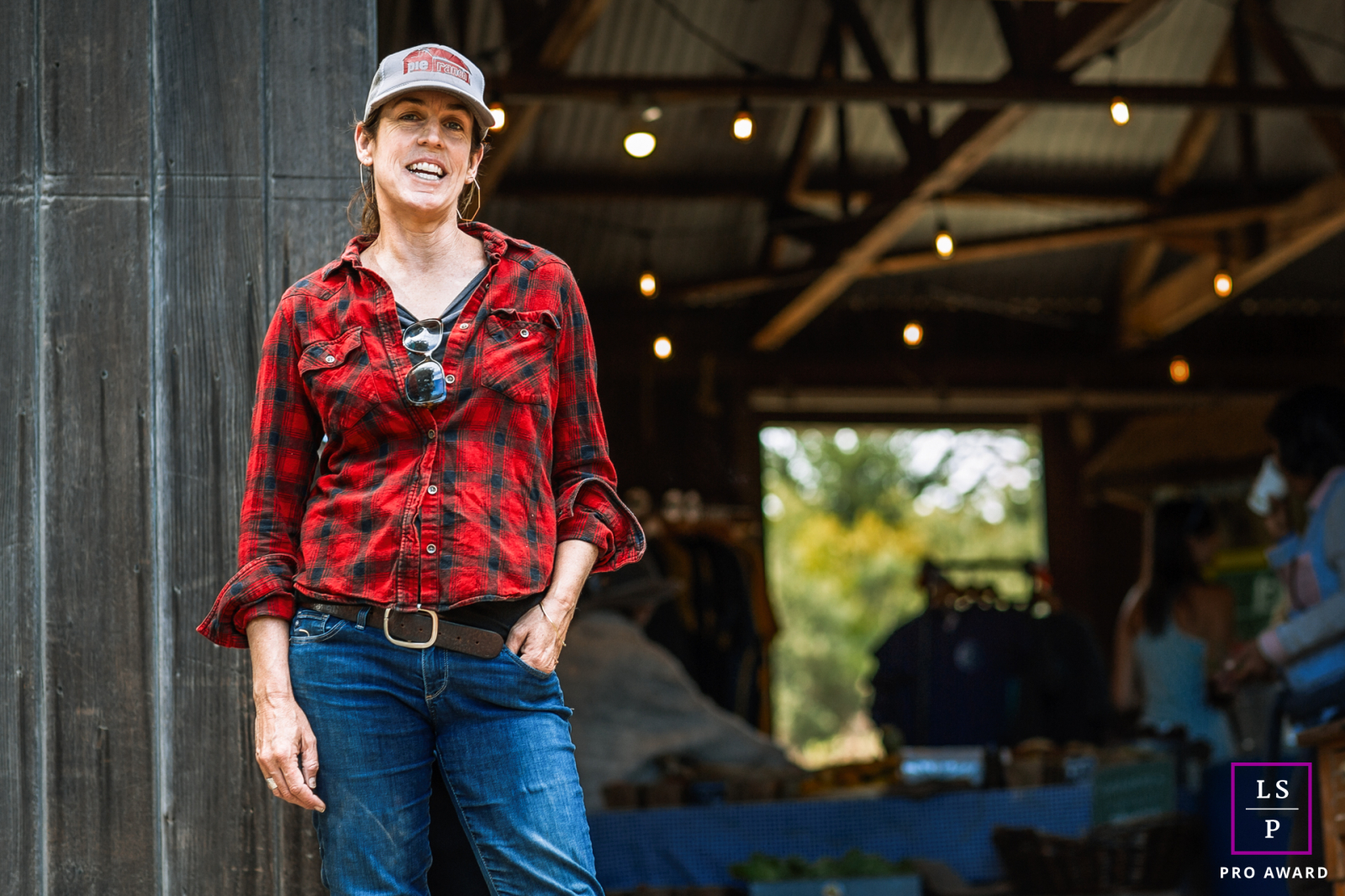 A photo of a Pescadero, CA woman in a hat, red shirt, and jeans standing in front of a farm building. It's a great day at Pie Ranch!