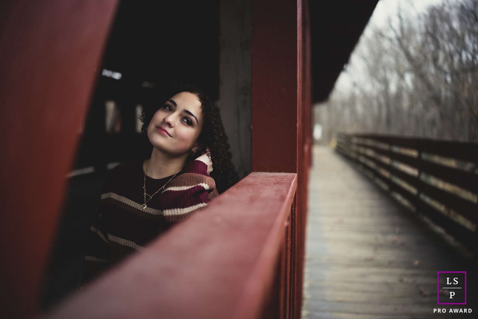 Uma jovem vibrante faz uma pausa em um sereno dia de outono em Mahomet, IL. Ela se inclina contra uma ponte rústica de madeira vermelha, adornada com delicados colares de ouro e um suéter listrado. Uma luz suave ilumina sua expressão calma e contente.