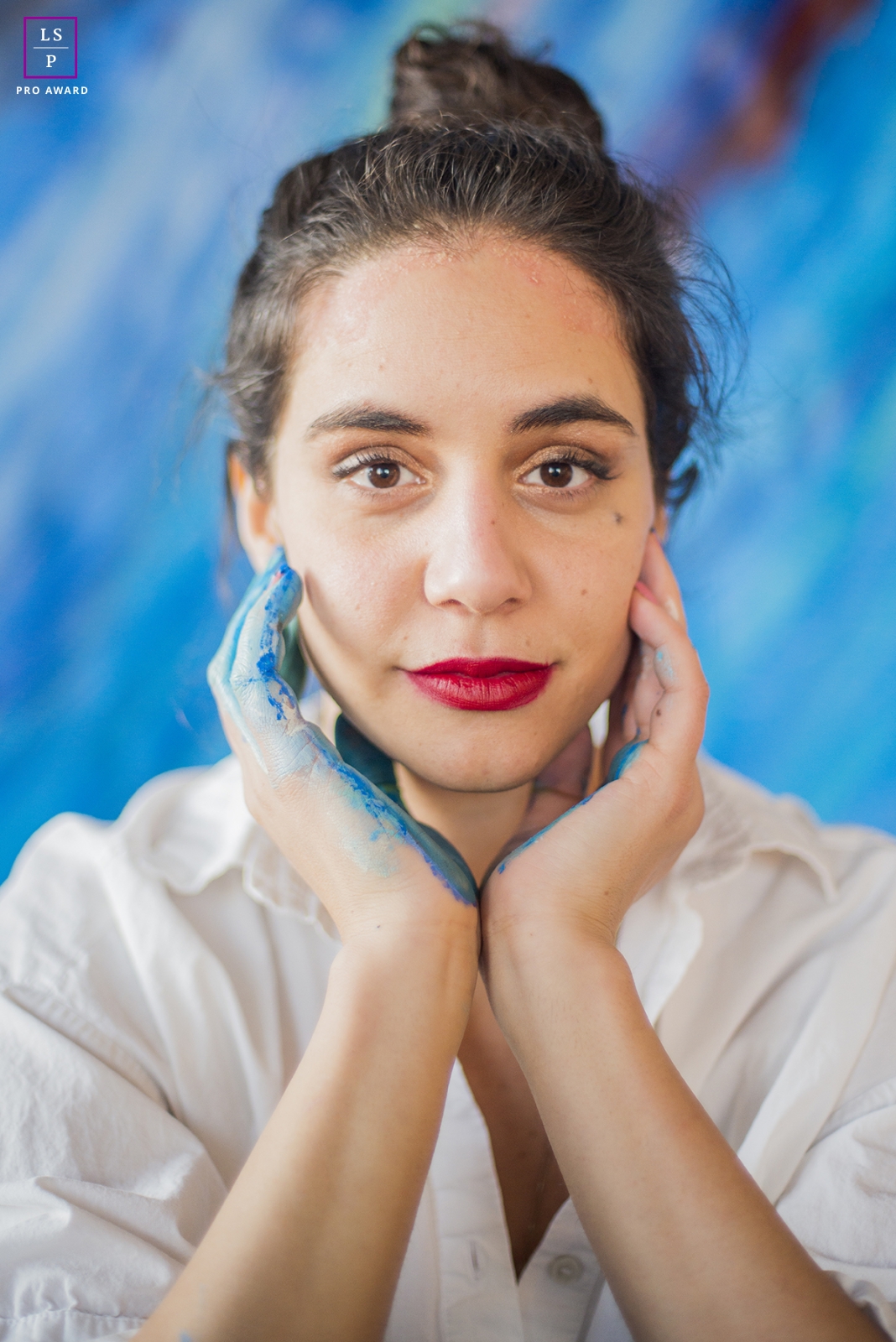 A lively and vibrant artist from the United States poses for a portrait, with blue paint on her hand near her face, against a matching blue background in New York.