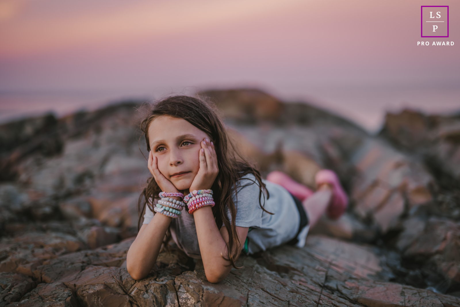 Retrato cativante do estilo de vida de uma jovem garota no Maine, EUA, perdida em pensamentos enquanto ela está deitada de bruços em pedras escarpadas da praia. Suas mãos sustentam sua cabeça, evocando uma sensação de tranquilidade e serenidade em meio à beleza natural da costa.