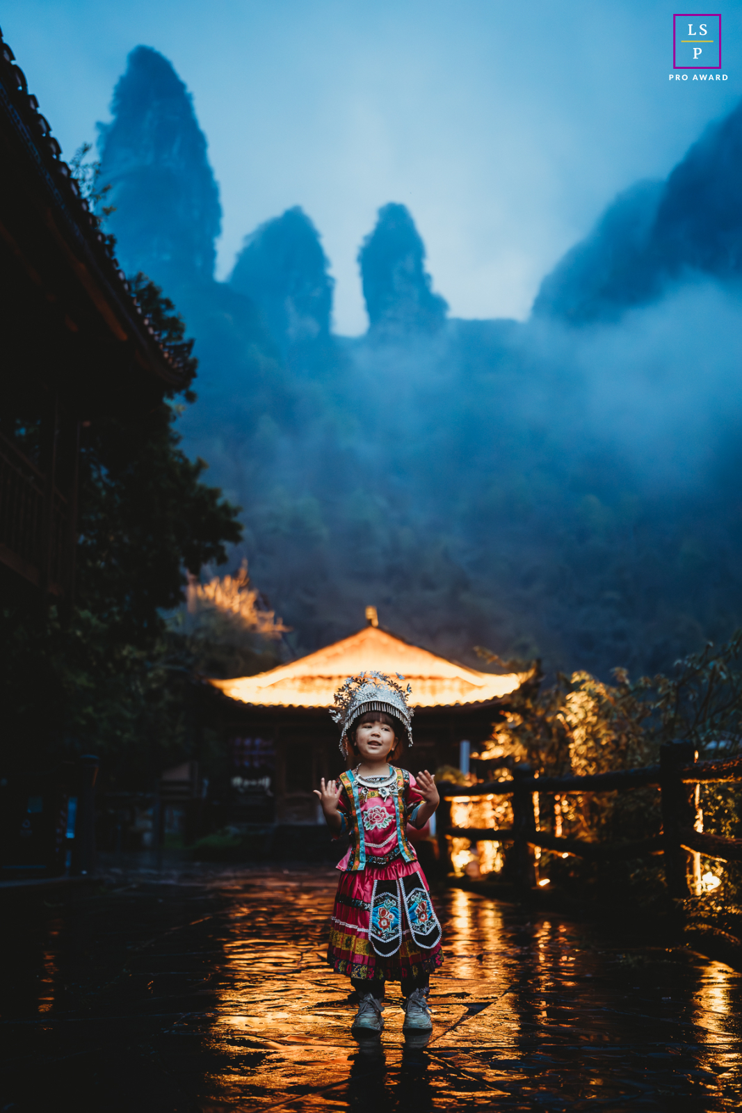 Uma garota usando um vestido tradicional de Yunnan posa contra um fundo azul vibrante, com neblina adicionando um elemento místico. Tirada no crepúsculo, cercada por árvores.