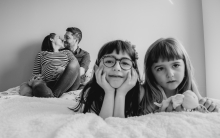 The black and white tones masterfully highlight a family in transition, layering the girls' curious foreground gazes against their parents' quiet, expectant embrace. It is a witty, heartwarming study of home.
