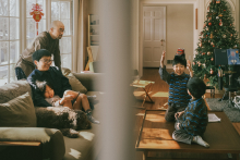 Seen through the divided doorway in this New Jersey home, the family unfolds in two simultaneous scenes of play and quiet rest. The layered framing and warm holiday light give the photograph depth, intimacy, and beautifully observed everyday storytelling.