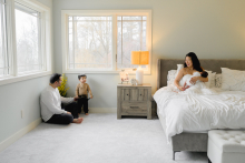 In NYC, the quiet rhythm of a growing family unfolds across the softly lit room as a mother cradles her newborn while father and son share a gentle moment nearby. The wide composition and calm tones give the portrait warmth, balance, and intimacy.
