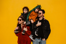 A family shares a playful embrace with two children perched on their parents’ shoulders against a bright yellow wall in Saint-Étienne, France.