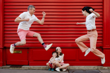 Parents leap playfully above their two seated children, creating a lively family scene against a bright red storefront in Paris.