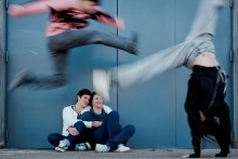 Two women sit together watching their teenagers leap and tumble playfully through the frame during a lively family session in Paris.