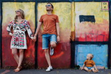 Retrato de família nas vibrantes ruas de Paris, cercado por um muro colorido adornado com grafite e obras de arte. Os pais estão de pé, de mãos dadas, com seu filho pequeno curioso sentado no chão, capturando a essência da vida urbana.