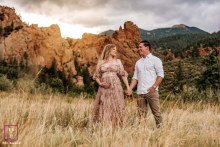 In Colorado Springs, CO, a pregnant mother stands hand in hand with her husband amidst tall grass, with the striking red rocks as their backdrop. The scene captures their united joy and anticipation.