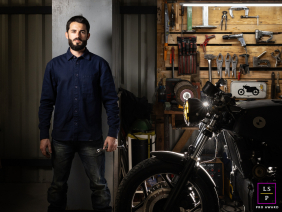 A motorcycle mechanic posing in Ruleshaker garage, Peyrehorade, Landes, France. The mechanic is focused on repairing a bike, surrounded by various tools and workshop equipment.