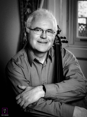 Cellist Rests Beside Instrument In Calm, Intimate Light Filled Black And White Braeside Portrait In Braeside, the cellist rests beside his instrument with the calm expression of someone who has spent years listening closely to music and silence alike. The black and white tones, soft window light, and intimate framing give the portrait timeless warmth