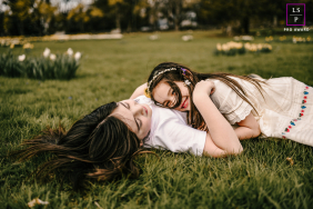 At Irwin Park, the eldest girl lies on her back on the grass while her younger sister hugs her, facing her. Their joyful expressions capture the warmth and affection of sibling love outdoors.